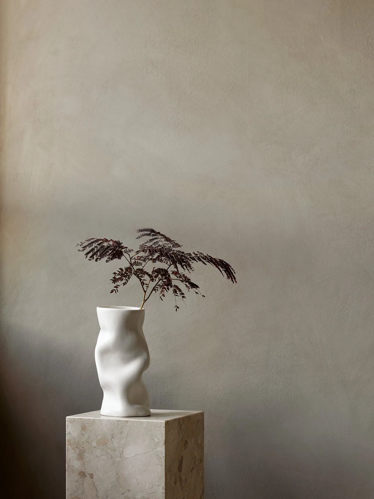 The sculptural white Collapse Vase holding a dark-leafed branch, styled on a square marble Plinth Pedestal.