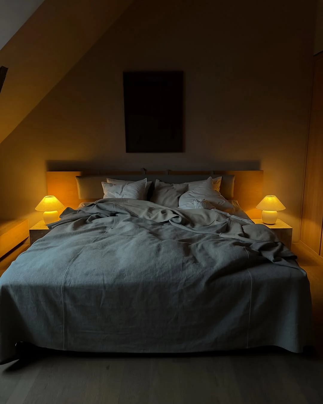 Minimal bedroom with king bed in grey linen, wooden headboard, two warm-glow ceramic bedside lamps and rumpled pillows and